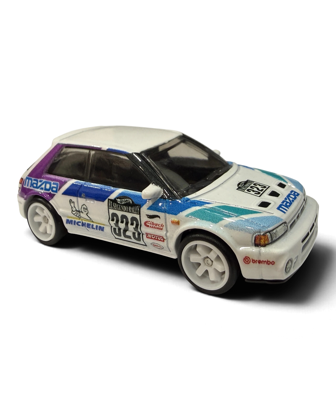VaultifyUK Hot Wheels Mazda 323 GTR die-cast car in white rally livery with purple and green graphics and white wheels.